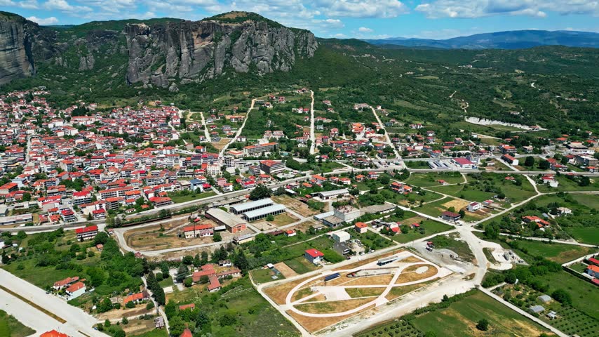 Aerial view of the town of Trikala with the dramatic Meteora rock formations in the background, surrounded by lush green fields and roads in Trikala, Greece.