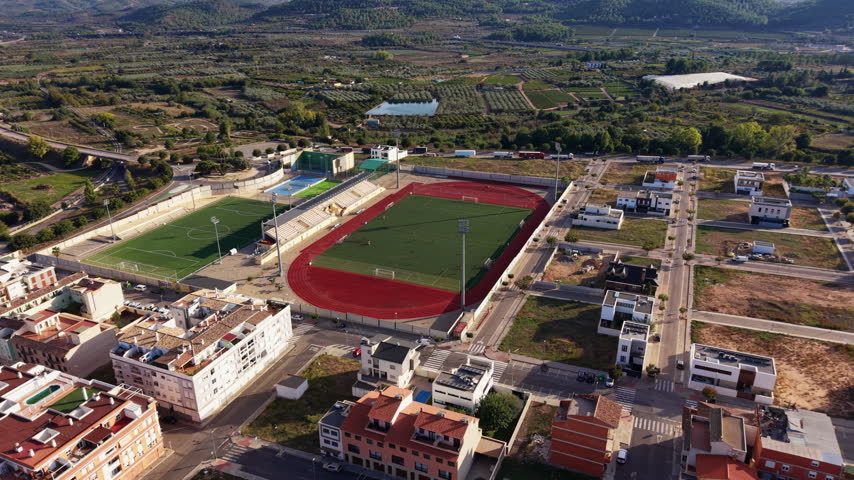 Elevated view of a football field, athletics track and residential buildings beside orchards and rural hills in a Mediterranean town