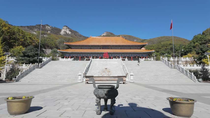 Forward moving view toward a traditional Chinese Buddhist temple hall across a wide stone plaza with an incense burner, mountains behind, autumn, ancient temple site in Dalian