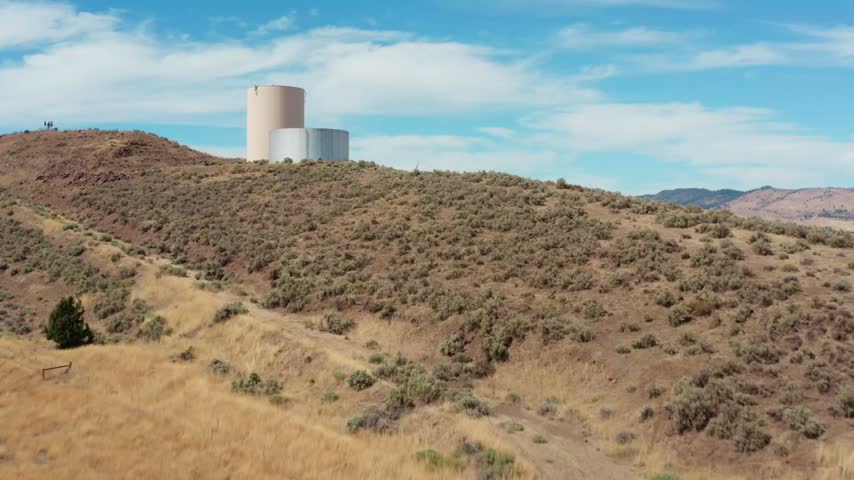 Drone rising over a sagebrush hilltop past a water tank, revealing a long freight rail yard full of railcars, the Klamath River and Lake Ewauna winding toward town, and dry mountains under a bright bl