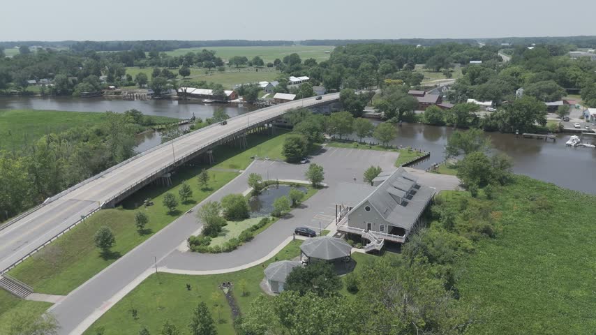Aerial flight past a highway bridge, a riverfront building and parking lot beside green tidal marsh along the Choptank River, Denton, Maryland, USA.