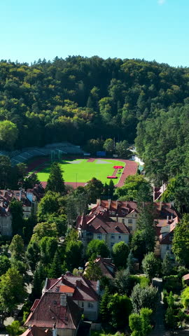 A highangle birds eye view of a sports stadium with a red running track and green field nestled between a dense forest hill and traditional European suburban architecture.