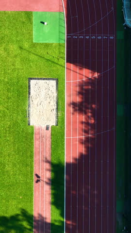 Topdown aerial perspective of a red synthetic running track, green grass field, and long jump sand pit at a sports stadium during golden hour.