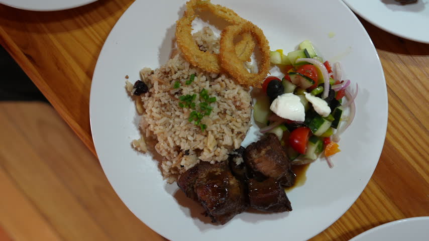 Horizontal close-up shot of a Colombian meal featuring seasoned rice, braised meat, crispy onion rings, and colorful fresh salad on a white plate, shot in warm indoor light.
