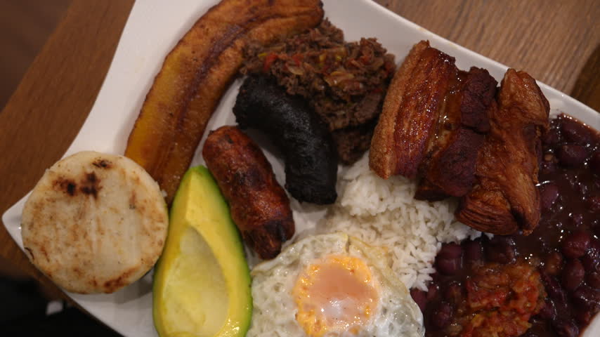 Vivid close-up shot of a traditional Colombian Bandeja Paisa with rice, fried egg, beans, plantain, avocado, chorizo, arepa, pork belly, and morcilla on a white plate under warm lighting.