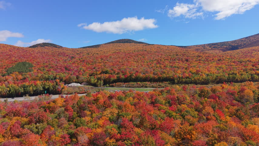 Highway with traffic surrounded by vibrant red and orange fall foliage and mountains in White Mountains, New Hampshire. Aerial forward