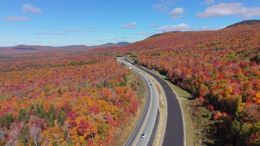 Highway with cars driving through vast woods with vibrant fall foliage towards mountains in White Mountains, New Hampshire. Aerial forward