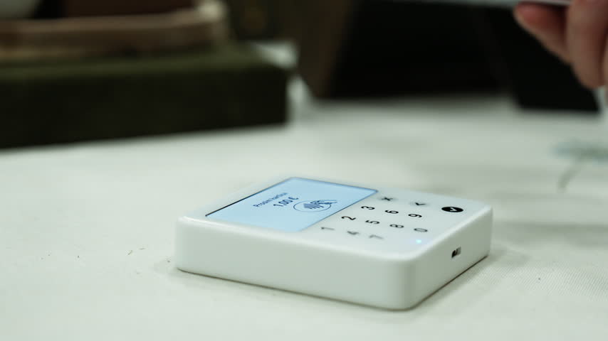 A hand holds a payment card over a small wireless reader on a table. The screen processes the contactless transaction and displays a confirmation.