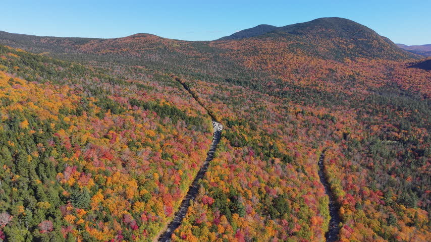 Highway cutting through vast woods with vibrant fall foliage towards distant mountains in White Mountains, New Hampshire. Aerial forward