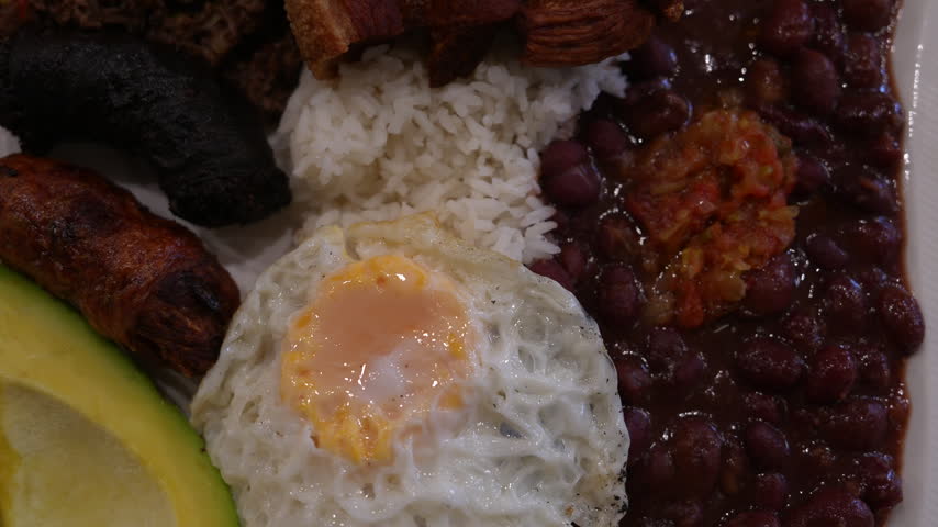 Detailed close-up shot of a Colombian bandeja paisa plate featuring fried egg, red beans, rice, sausage, plantain, avocado, and pork, in natural indoor lighting. Rich colors and textures.