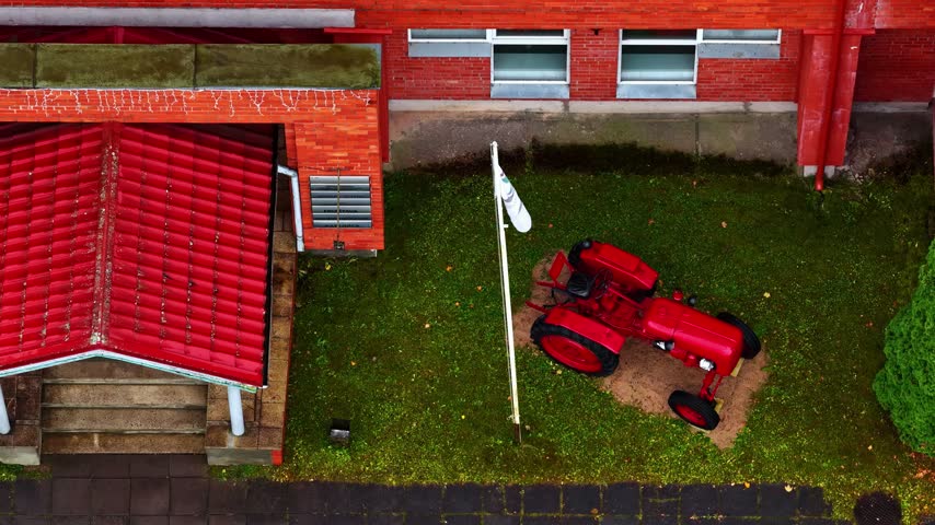 Aerial view of a red vintage tractor parked on a green lawn next to a building with a red roof in Priekuli, Cesis Municipality, Latvia.
