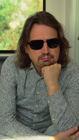 Adult man wearing sunglasses rests his chin on his hand while sitting in a metro train as trees and buildings move past the window in Amsterdam.