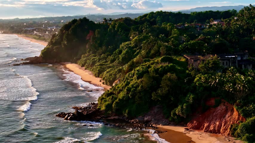 Aerial view of Madiha Beach with lush green cliffs, sandy shoreline, and waves crashing against the rocks in Matara, Southern Province, Sri Lanka.