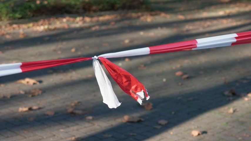 Red and white barrier tape tied to a metal rail over a paved path with fallen leaves