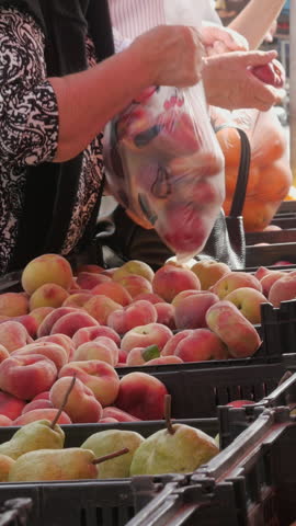 Street Market Fruit Stall Shoppers Selecting Fresh Peaches and Kiwis. Side view shot captures shoppers purchasing fresh fruit at a street market stall, with a customer holding a packed plastic bag over crates filled with flat peaches, pears, and kiwis.