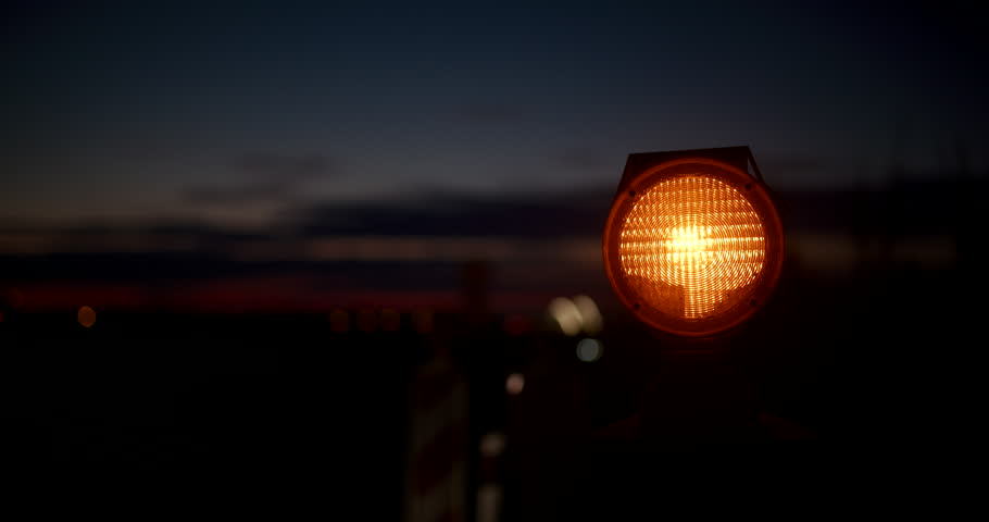 Glowing amber warning light on a roadside at dusk, with blurred red taillights and a dark twilight sky with clouds in the background, copy space on the left. 4K selective focus.