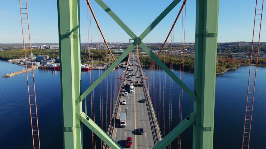 An aerial view follows cars crossing the A. Murray MacKay Bridge above blue Halifax Harbour in Halifax, Nova Scotia, Canada, with green steel towers framing the roadway.
