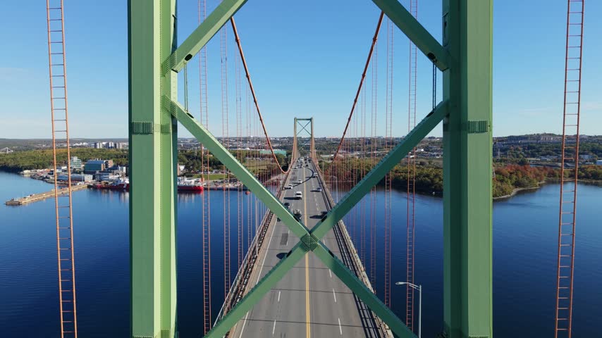 An aerial view follows the A. Murray MacKay Bridge over blue Halifax Harbour, with cars crossing between Halifax and Dartmouth beneath a bright daytime sky in Nova Scotia, Canada.