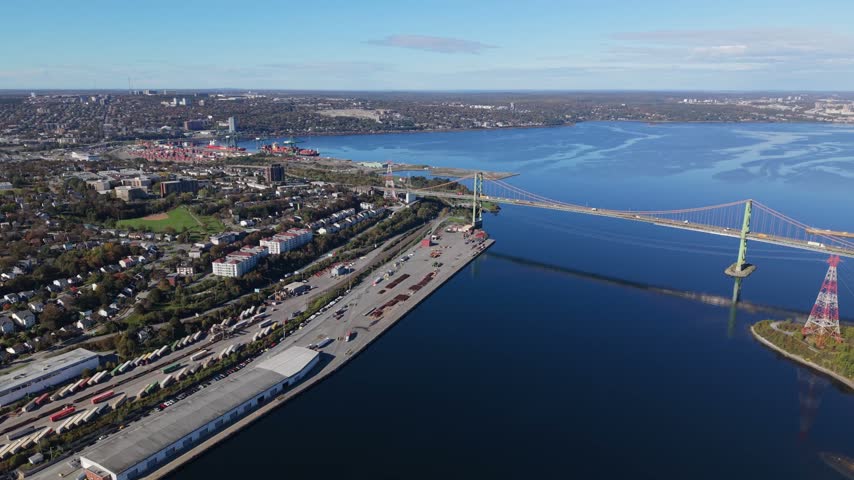 A steady aerial view reveals the A. Murray MacKay Bridge, shoreline roads, and Halifax waterfront beneath a bright daytime sky in Nova Scotia, Canada.