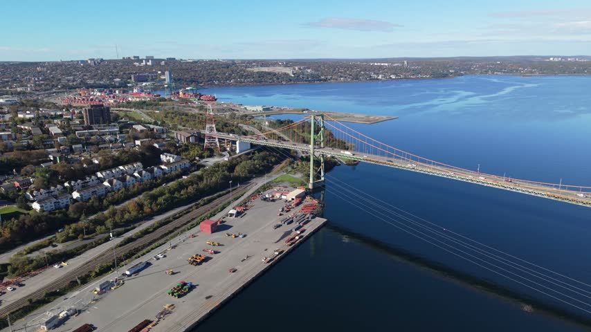 A calm aerial view follows the A. Murray MacKay Bridge across Halifax Harbour in Halifax, Nova Scotia, Canada, with traffic moving between the green shoreline and blue water in daylight.