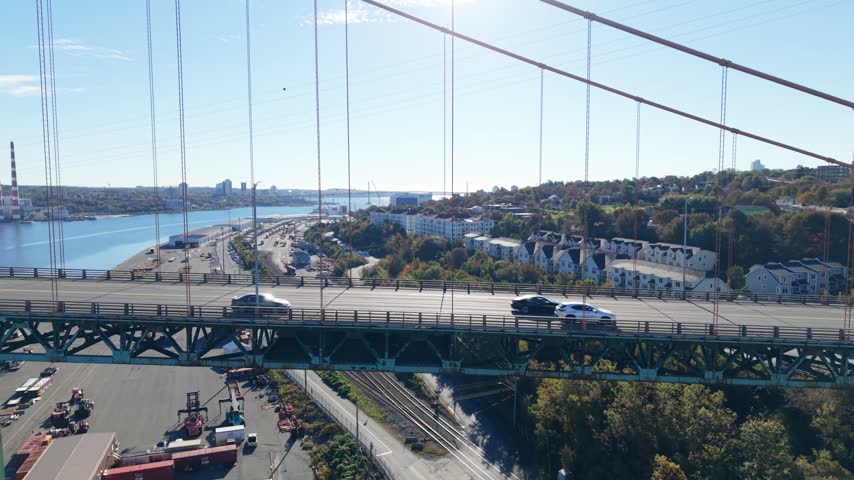 Vehicles travel along a Halifax highway interchange beneath a green suspension bridge, with the harbor and waterfront visible in daylight in Nova Scotia, Canada.
