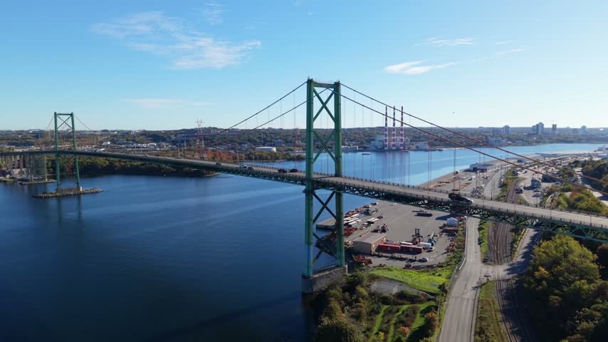 An aerial view of traffic moving along a Halifax highway beside the Macdonald Bridge, spanning blue water under a bright daytime sky in Nova Scotia, Canada.
