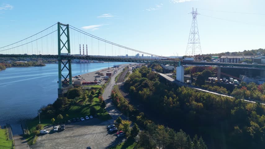 An aerial view follows traffic along a Halifax highway interchange beside a suspension bridge, with blue water, leafy shores, and city buildings in Nova Scotia, Canada, by day.