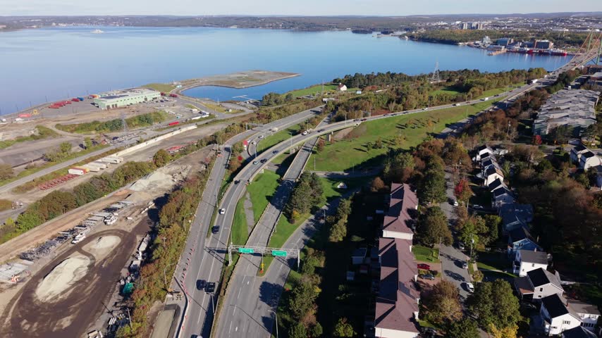 An aerial view follows traffic along a broad highway interchange in Halifax, Nova Scotia, with green space, nearby homes, and blue coastal water in the distance.