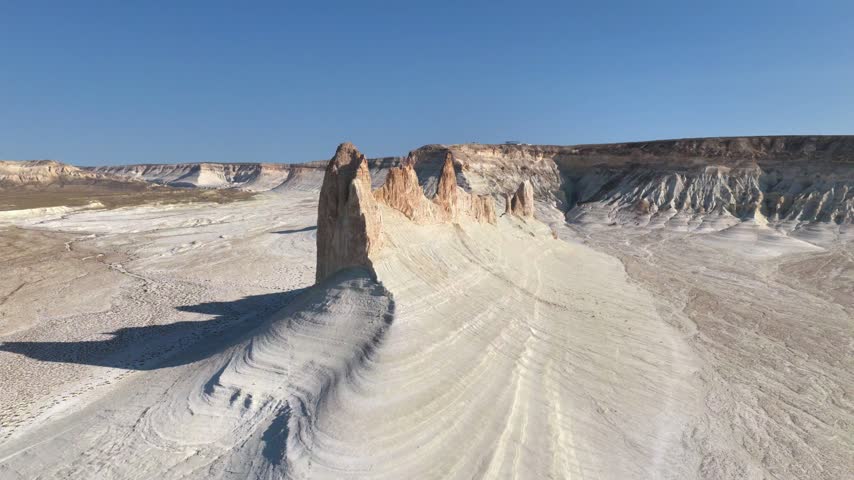 The drone flies beside Bozzhyra Dragon Crest as its pale sculpted ridge gives way to warm orange pinnacles, backed by layered escarpments and clear sky.