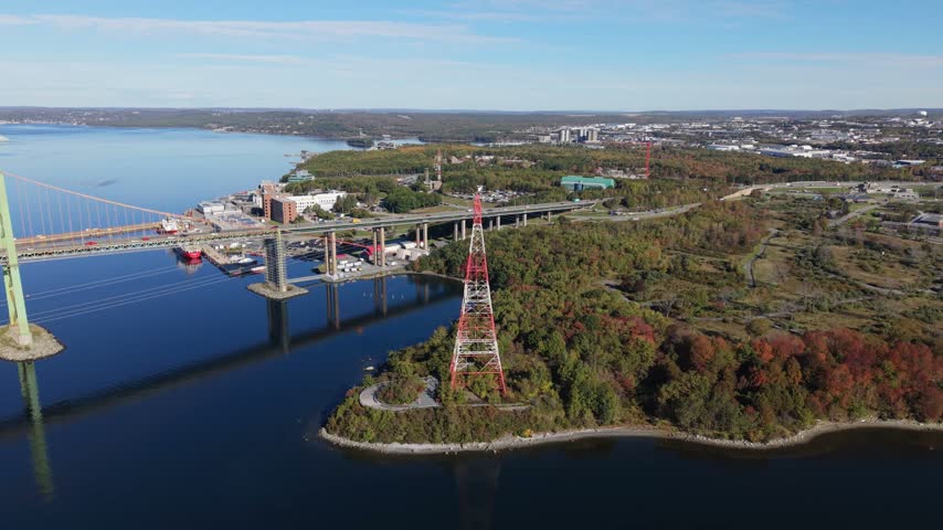 An aerial view follows Nova Scotia Power transmission towers beside calm water and autumn-colored trees, with roads, buildings, and the Nova Scotia landscape visible in daylight.
