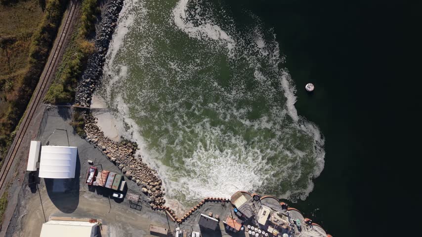 An aerial view of Nova Scotia Power plant pipes beside a rocky shoreline in Canada, where water churns and swirls in bright foam during the day.