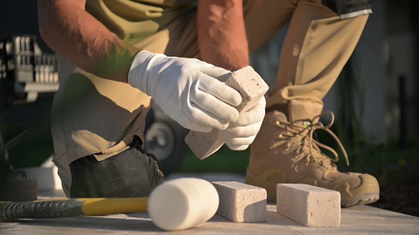 A worker kneels beside stacked paving blocks and uses a mallet to adjust the patio surface.