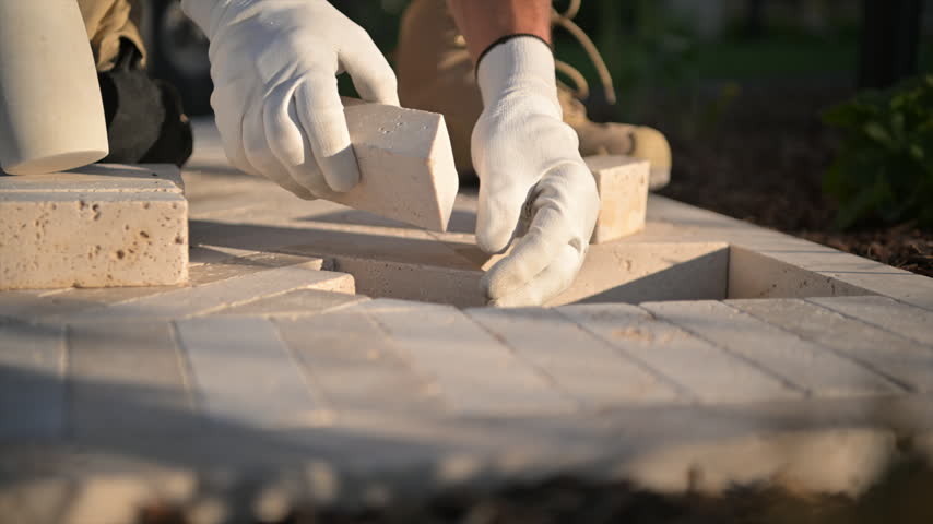 Low ground view of hands fitting a rectangular block into a herringbone patio and tapping it into alignment.