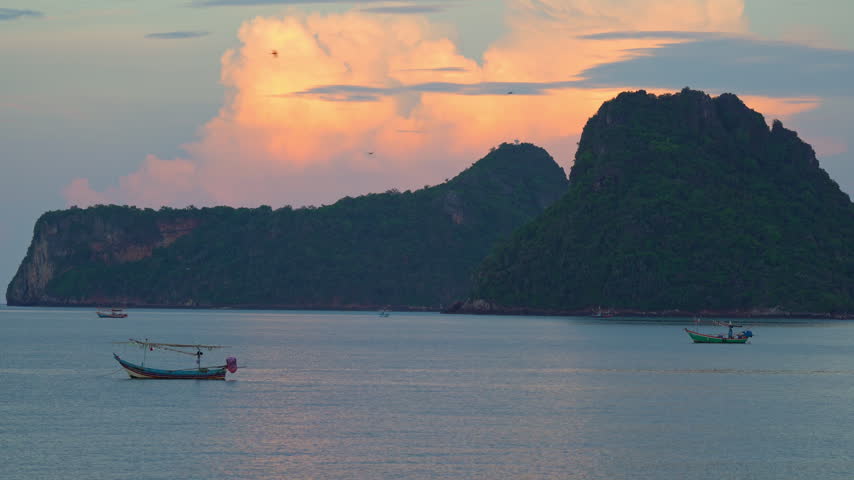 Static shot of traditional wooden fishing boats anchored in calm sea water at Prachuap Khiri Khan Bay with birds flying across a golden sunset sky, Thailand.