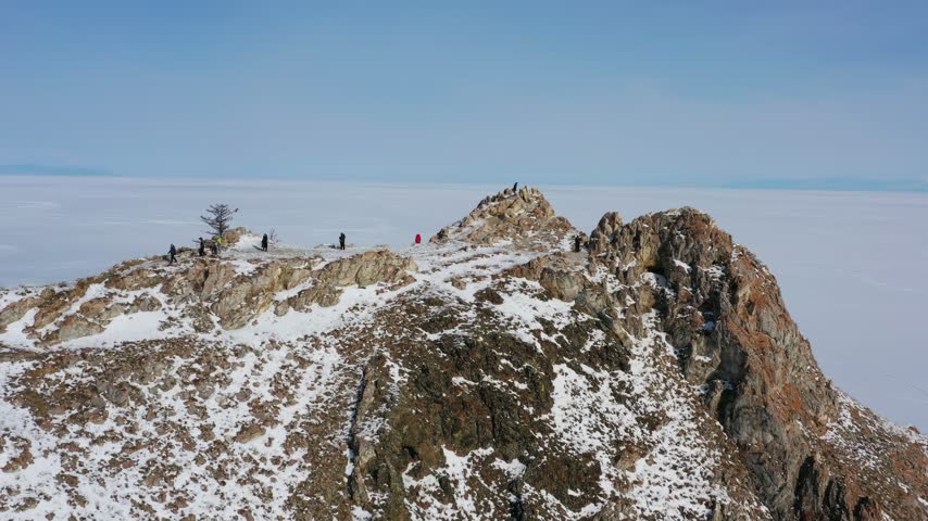 Aerial view of Lake Baikal snowy mountain peak with people standing on the rocky summit above a sea of clouds in Russia.