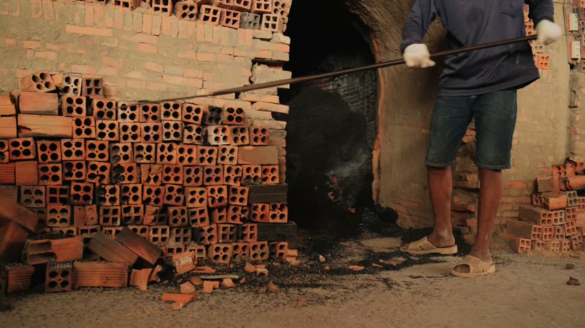 A worker tends a brick kiln with a long pole beside stacks of hollow clay bricks, with soot and broken bricks on the dusty ground in Mang Thit, Vietnam.