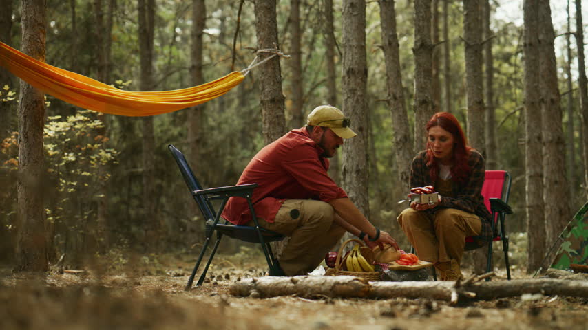 Couple shares food while camping at a pine forest campsite, sitting in folding chairs beside a hammock.