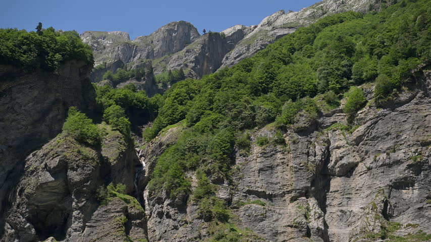 Dense green foliage covers the tops of layered cliffs in a sunny mountain landscape in the French Alps.