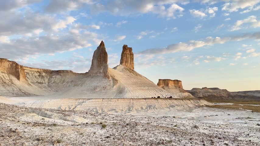 A low aerial pan moves from the Bozzhyra Fangs toward a small desert camp and parked vehicles beneath scattered clouds at sunrise.