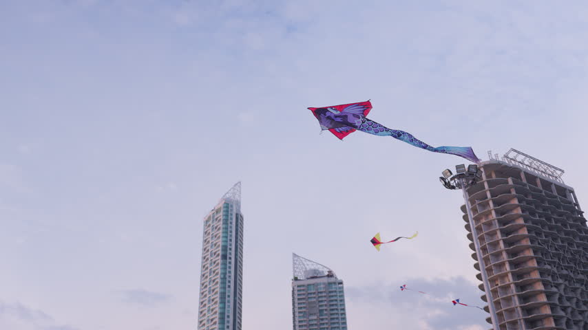 A large dragon kite soars gracefully over a modern city, its playful flight contrasting with the urban landscape of skyscrapers and a building under construction.