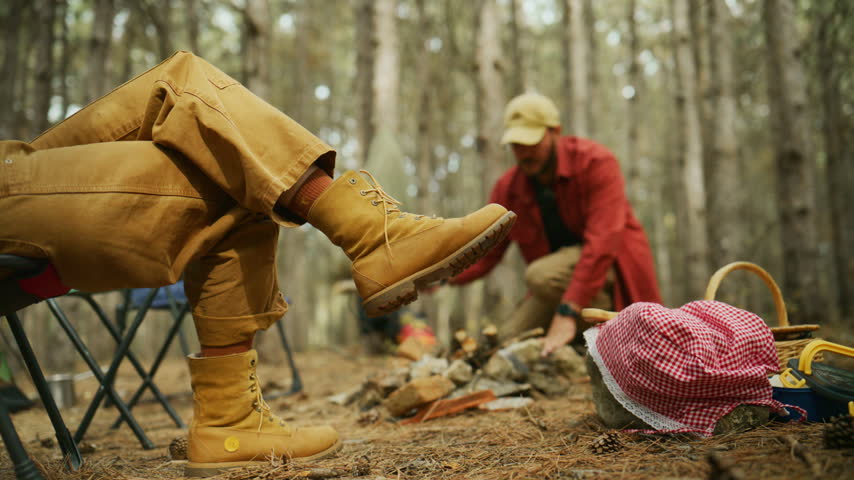Camper sits in a chair while a companion tends a campfire at a forest campsite, with outdoor camping and leisure.