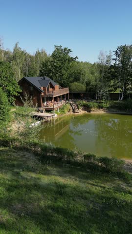 Camera movement reveals a calm green pond, wooden house, dock, grassy bank, and surrounding trees under a clear blue sky