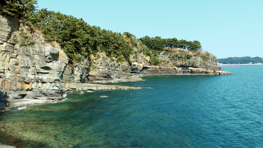 Aerial view of a traditional Korean pavilion on a rocky coastal cliff with pine trees overlooking blue sea at Goseong, Gyeongsangnam-do, South Korea.