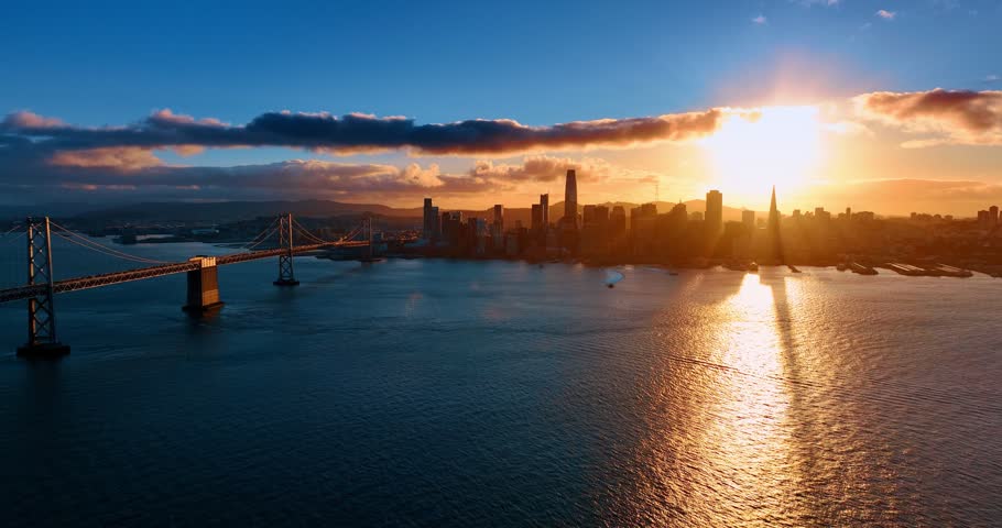 Skyline of San Francisco, California, USA set against orange sky at sunset. Two vessels approach by the waterscape colored by the setting sun.
