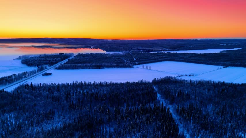 Spectacular orange sky over the vast woods. Drone footage approaching the snowy field at sunset.