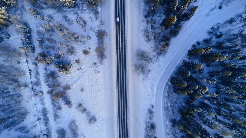 Few cars ride by the straight highway crossing the woodlands. Snow covers the land, trees and houses roofs. Top view.