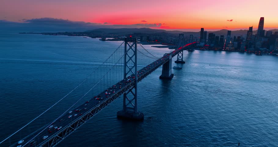 Approaching the support of the Oakland Bay Bridge with red light flashing on. Multiple cars drive by the bridge at sunset. San Francisco, California, USA.