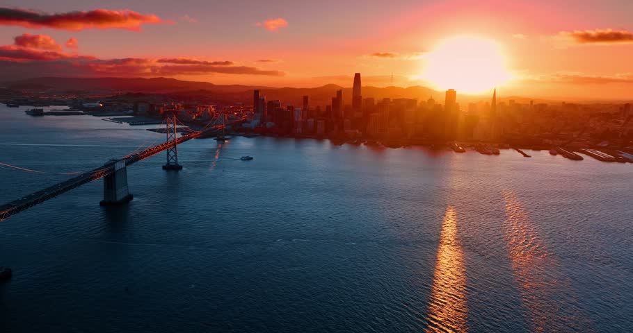 View on San Francisco skyline lit with golden light of sunset. Numerous cars move by the Oakland Bay Bridge in hectic traffic. Aerial perspective.