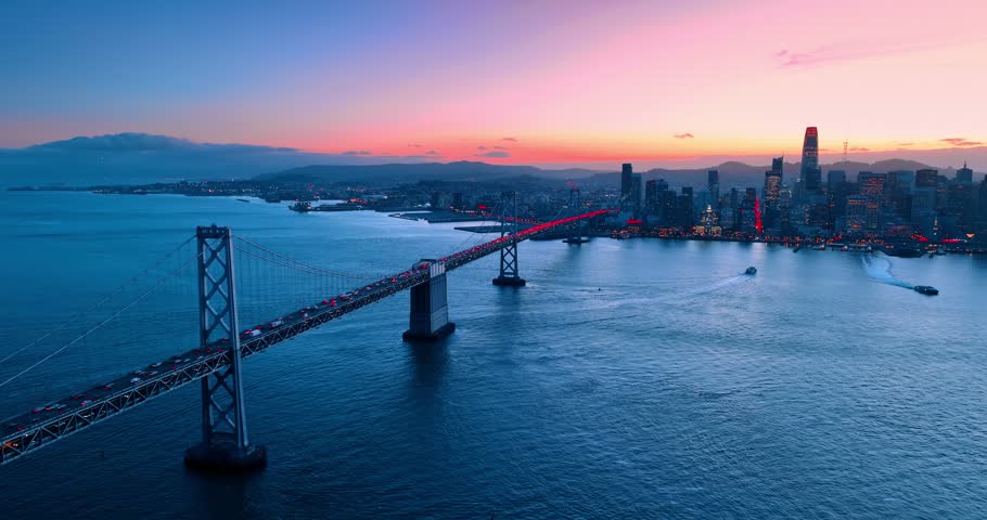 Huge amount of cars move by the Oakland Bay Bridge to and from downtown San Francisco, California, USA. City skyline against pink sky at sunset in the backdrop.