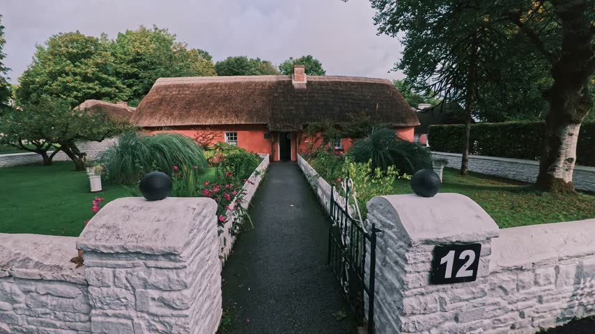 Horizontal stock video footage of a traditional pink-painted Irish cottage with a thatched roof, white picket fence, and a scenic garden pathway leading to the front door in rural Ireland.